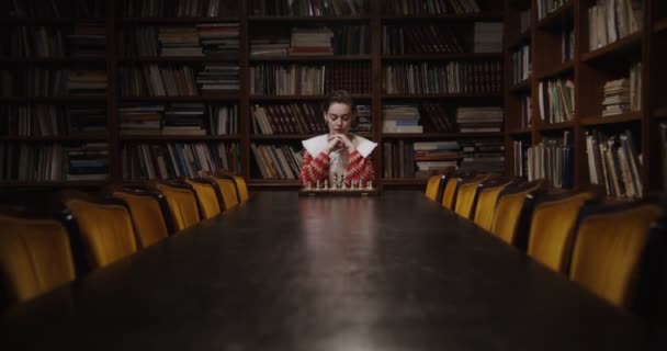 Una joven juega una partida de ajedrez sola, sentada en una mesa larga en la biblioteca — Vídeos de Stock
