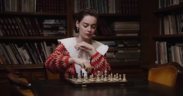 Uma jovem joga um jogo de xadrez sozinha, sentada em uma longa mesa na biblioteca — Vídeo de Stock