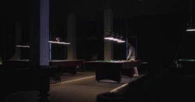 Bir adam bilardo kulübünde loş ışıkların altında tek başına bilardo oynuyor.
