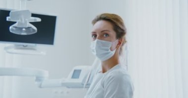 Bir kadın diş hekimi dişçide otururken doğrudan kameraya bakar.