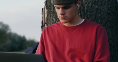 Genç bir adam dizüstü bilgisayar kullanıyor, açık havada bir ağacın altında oturuyor.