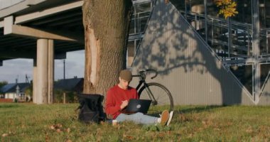 Adamın biri tablet ve dizüstü bilgisayar kullanıyor. Ağacın altında oturuyor.