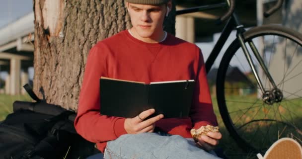 Seorang pemuda membaca sebuah buku dan makan sandwich, duduk di tanah di bawah pohon — Stok Video