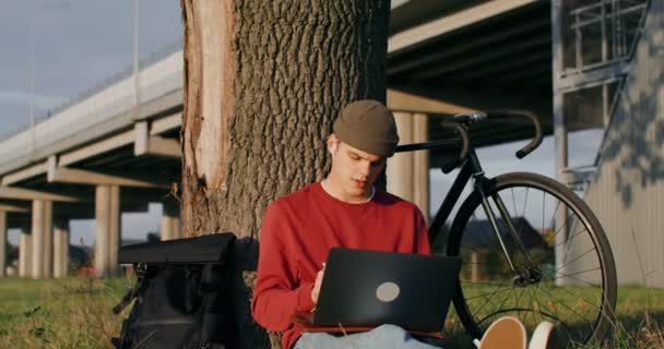 Ένας άνθρωπος χρησιμοποιεί ένα tablet γραφικών και ένα φορητό υπολογιστή, κάθεται στο έδαφος κάτω από ένα δέντρο — Αρχείο Βίντεο