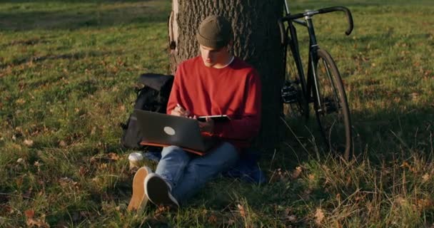 Um homem usa um tablet gráfico e um laptop, sentado no chão sob uma árvore — Vídeo de Stock