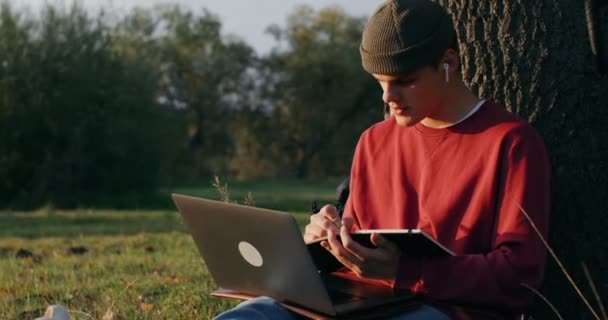 Een jongeman gebruikt een laptop en schrijft in een notitieboekje terwijl hij onder een boom zit — Stockvideo