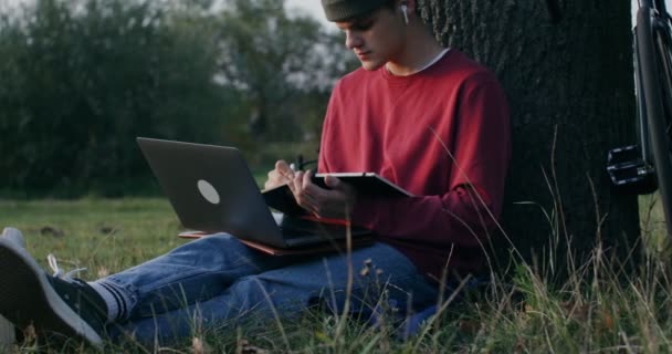 Un giovane uomo usa un portatile e scrive in un quaderno mentre è seduto sotto un albero — Video Stock