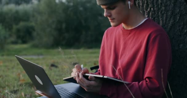 Een jongeman gebruikt een laptop en schrijft in een notitieboekje terwijl hij onder een boom zit — Stockvideo