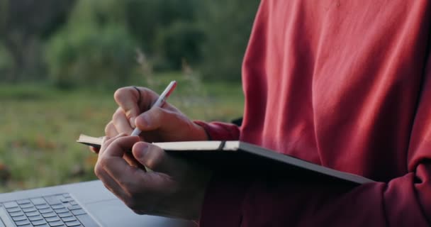 Młody człowiek robi notatki w notatniku, używając laptopa, siedząc na ziemi. — Wideo stockowe