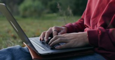 Genç bir adam dışarıda bir ağacın altında çimlerin üzerinde dizüstü bilgisayar kullanıyor..