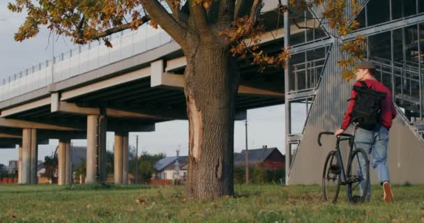 Een man leidt een fiets in zijn handen, stopt onder een boom en gaat zitten. — Stockvideo