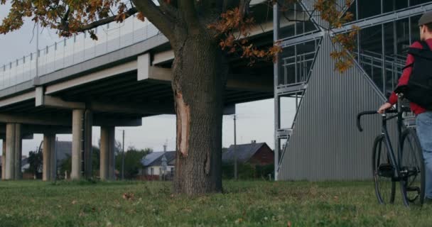 Een man leidt een fiets in zijn handen, stopt onder een boom en gaat zitten. — Stockvideo