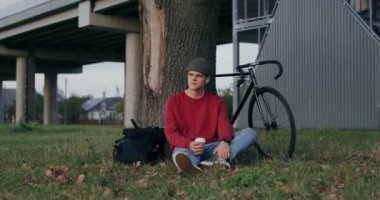 Genç bir adam bir ağacın altında yerde otururken kahve içer.