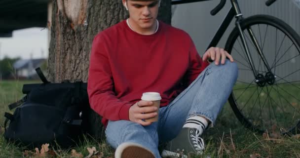 Молодой человек пьет кофе, сидя на земле под деревом — стоковое видео