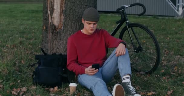 Un joven usa un teléfono mientras está sentado en el suelo bajo un árbol — Vídeos de Stock