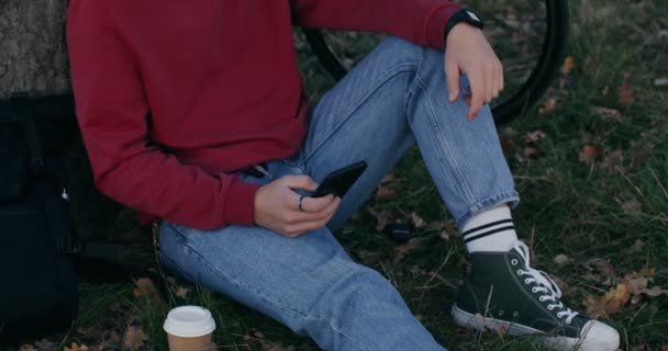 Молодой человек использует телефон, сидя на земле под деревом — стоковое видео