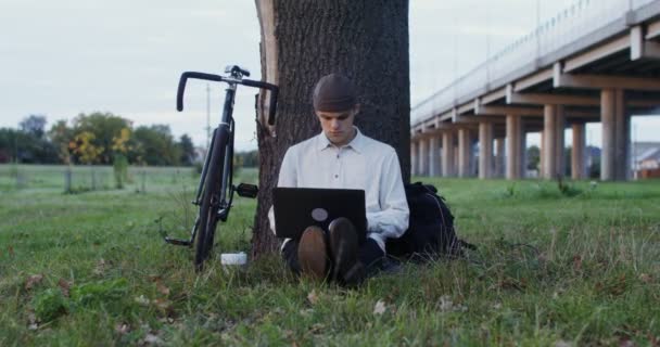 Un giovane uomo usa un computer portatile e beve caffè, seduto a terra sotto un albero — Video Stock