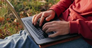 Genç bir adam dışarıda bir ağacın altında çimlerin üzerinde dizüstü bilgisayar kullanıyor..