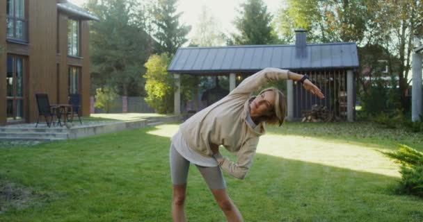 Une jeune femme en vêtements de sport fait des exercices sur la pelouse près d'un chalet moderne — Video