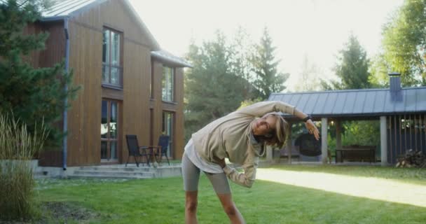 Une jeune femme en vêtements de sport fait des exercices sur la pelouse près d'un chalet moderne — Video