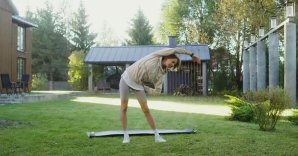 Mladá žena ve sportovním oblečení cvičí na trávníku v blízkosti moderní chaty — Stock video
