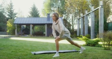 Spor giyimli genç bir kadın modern bir kır evinin yakınındaki çimenlikte egzersiz yapıyor.