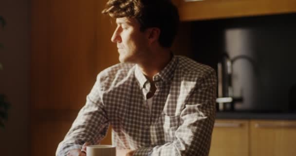 Mladý muž pije horký čaj nebo kávu, zatímco sedí u stolu v kuchyni — Stock video