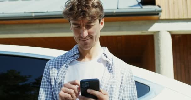Ένας νεαρός άνδρας χρησιμοποιεί ένα κινητό τηλέφωνο ενώ στέκεται στην πίσω αυλή ακουμπώντας σε ένα αυτοκίνητο — Αρχείο Βίντεο