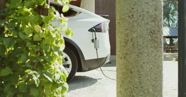 Un coche eléctrico con un cable de alimentación conectado a un cargador se encuentra en el patio trasero — Vídeos de Stock