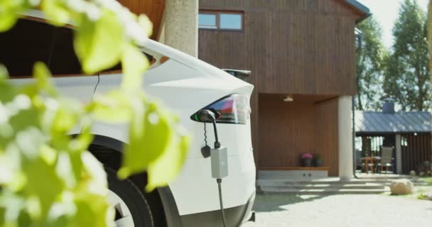 Un coche eléctrico con un cable de alimentación conectado a un cargador se encuentra en el patio trasero — Vídeo de stock