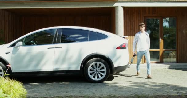 Um homem conecta um cabo de alimentação a um carro elétrico estacionado no quintal — Vídeo de Stock