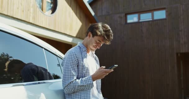 Un tânăr folosește un telefon mobil în timp ce stă în curtea din spate sprijinindu-se pe o mașină — Videoclip de stoc