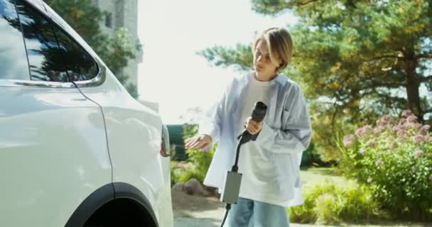 Schöne junge Frau verbindet Stromkabel mit weißem Elektroauto — Stockvideo