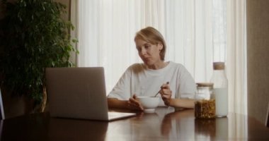 Genç bir kadın mısır gevreğini sütle yer, dizüstü bilgisayarın önünde oturur.