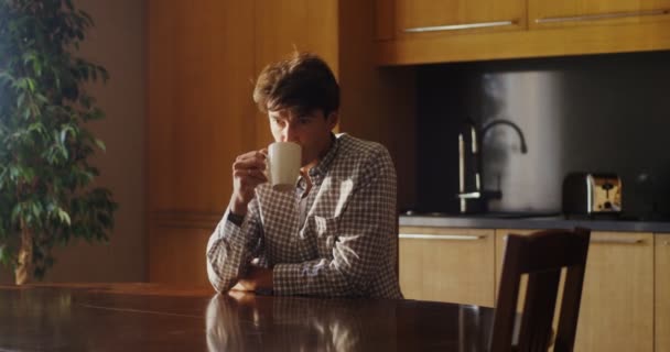A young man is drinking hot tea or coffee while sitting at a table in kitchen — Stock Video
