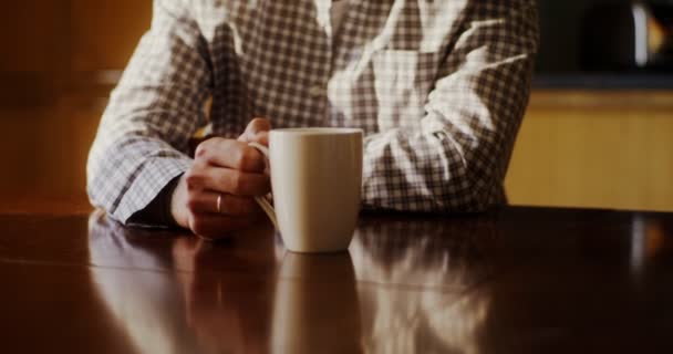 Een jongeman drinkt warme thee of koffie terwijl hij aan een tafel in de keuken zit — Stockvideo