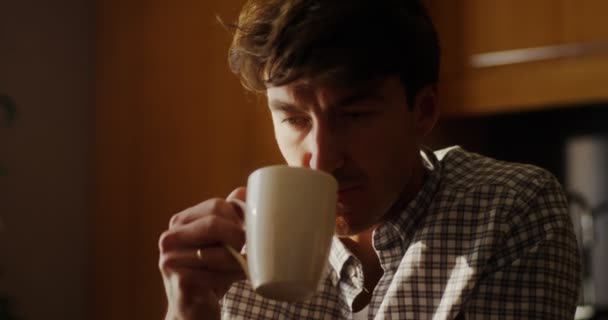 Ένας νεαρός άνδρας πίνει ζεστό τσάι ή καφέ, ενώ κάθεται σε ένα τραπέζι στην κουζίνα — Αρχείο Βίντεο