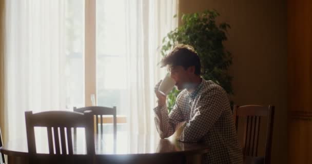 A young man is drinking hot tea or coffee while sitting at a table in kitchen — Stock Video