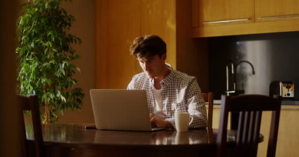 Ένας νεαρός άνδρας χρησιμοποιεί φορητό υπολογιστή και πίνει καφέ ενώ κάθεται σε ένα τραπέζι στην κουζίνα — Αρχείο Βίντεο