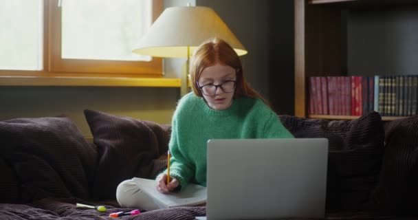 Uma menina faz anotações no caderno, sentada no sofá e olhando para um laptop — Vídeo de Stock