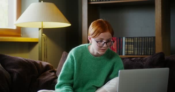 A red-haired girl with glasses starts video chat on a laptop, sitting on a sofa — Stock Video