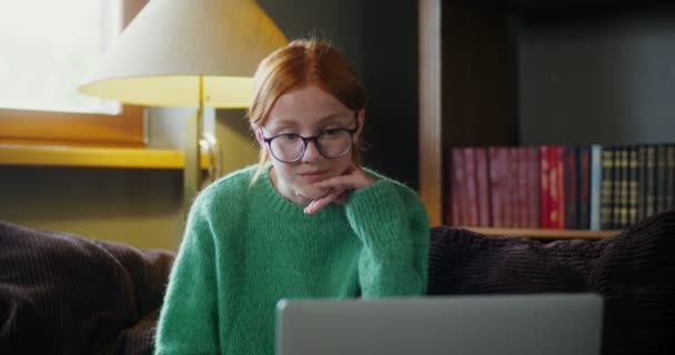 A red-haired girl with glasses starts video chat on a laptop, sitting on a sofa — Stock Video