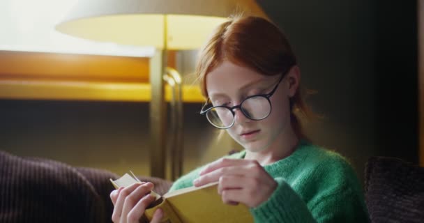 Egy szemüveges lány könyvet olvas álmos tekintettel, a kanapén ülve. — Stock videók