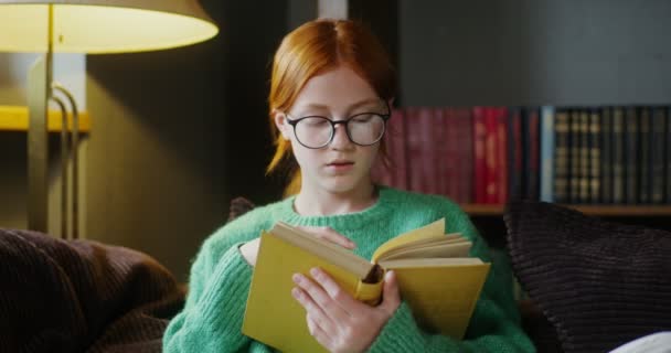 Ένα κορίτσι με γυαλιά που διαβάζει ένα βιβλίο με ένα ονειρεμένο βλέμμα, κάθεται στον καναπέ — Αρχείο Βίντεο