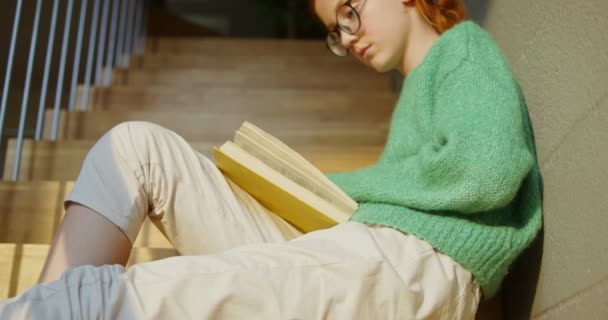 Una chica leyendo un libro atentamente mientras está sentada en las escaleras en el interior de una casa — Vídeos de Stock