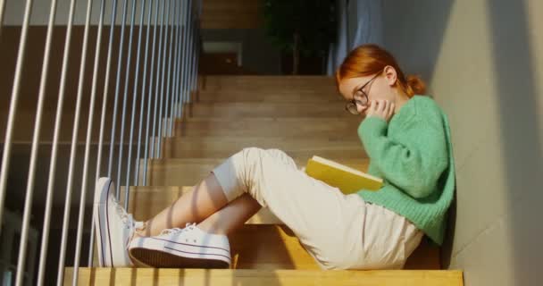 En flicka läser en bok intensivt när hon sitter på trappan i en heminredning — Stockvideo
