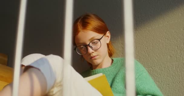 Una chica leyendo un libro atentamente mientras está sentada en las escaleras en el interior de una casa — Vídeos de Stock