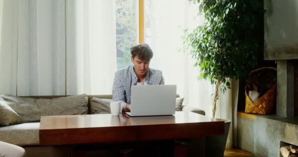 在现代室内，一个男人坐在咖啡桌旁的沙发上，一边用笔记本电脑 — 图库视频影像