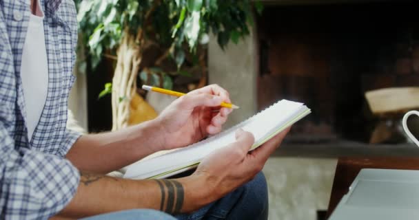 Un hombre toma notas en un cuaderno mientras está sentado cerca de un portátil abierto — Vídeos de Stock