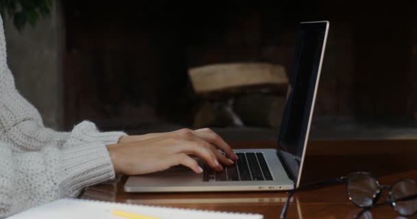 Μια γυναίκα δακτυλογραφεί σε ένα φορητό υπολογιστή κάθεται σε ένα τραπεζάκι δίπλα στο τζάκι — Αρχείο Βίντεο